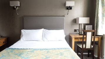 A hotel room with a large bed, two bedside lamps, a wooden nightstand, a desk with a chair, and a window with curtains.
