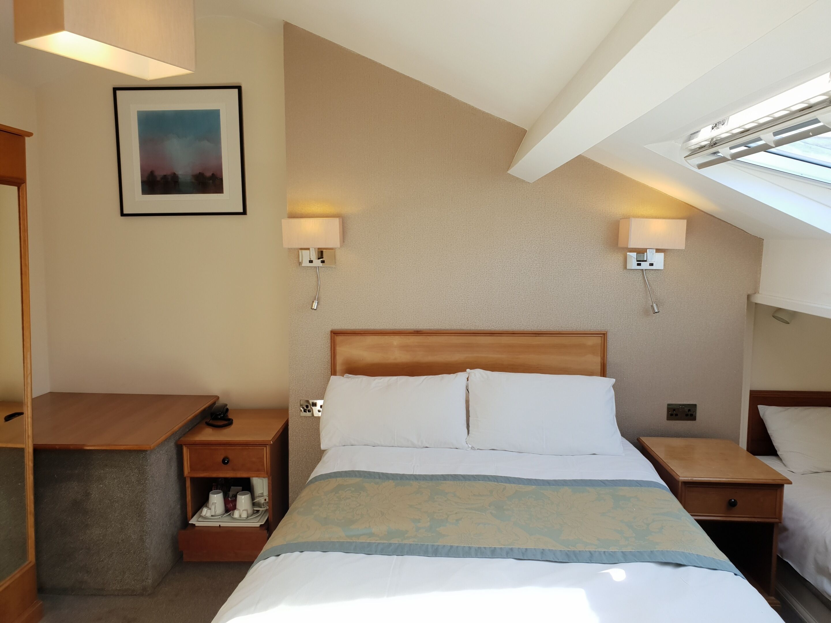 A hotel room with a bed, bedside tables, a desk, a lamp, a picture on the wall, and a skylight.