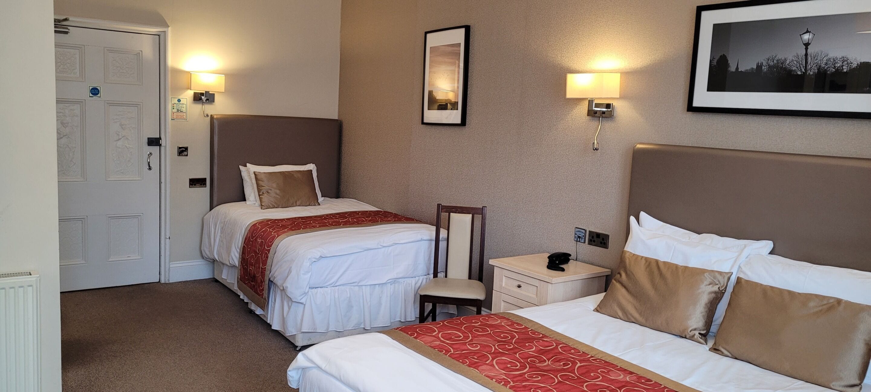 A hotel room with two beds, a nightstand, a chair, a mirror, and wall-mounted lights.