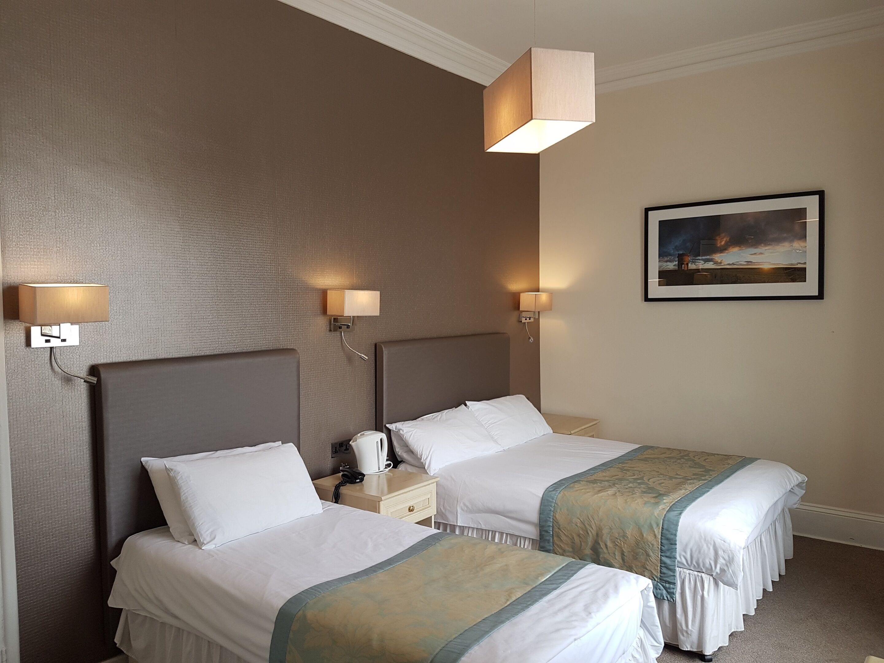 A hotel room with two beds, each with a headboard, bedside tables, and lamps. There is a framed picture on the wall.