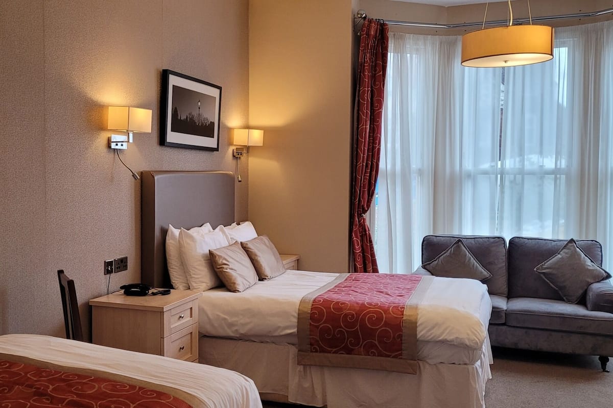 Ein Hotelzimmer mit zwei Betten, einem Sofa, einem Schreibtisch und einem großen Fenster mit Vorhängen.