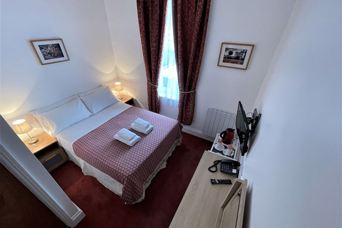 Quarto de hotel com cama, mesa de cabeceira com abajur, escrivaninha com computador e duas fotografias emolduradas nas paredes.