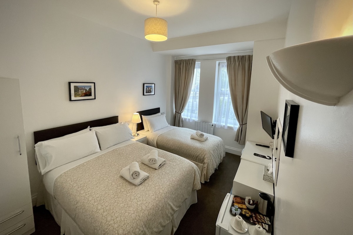 Quarto de hotel com duas camas, uma mesa de escritório com computador, uma luminária e uma janela com cortinas.