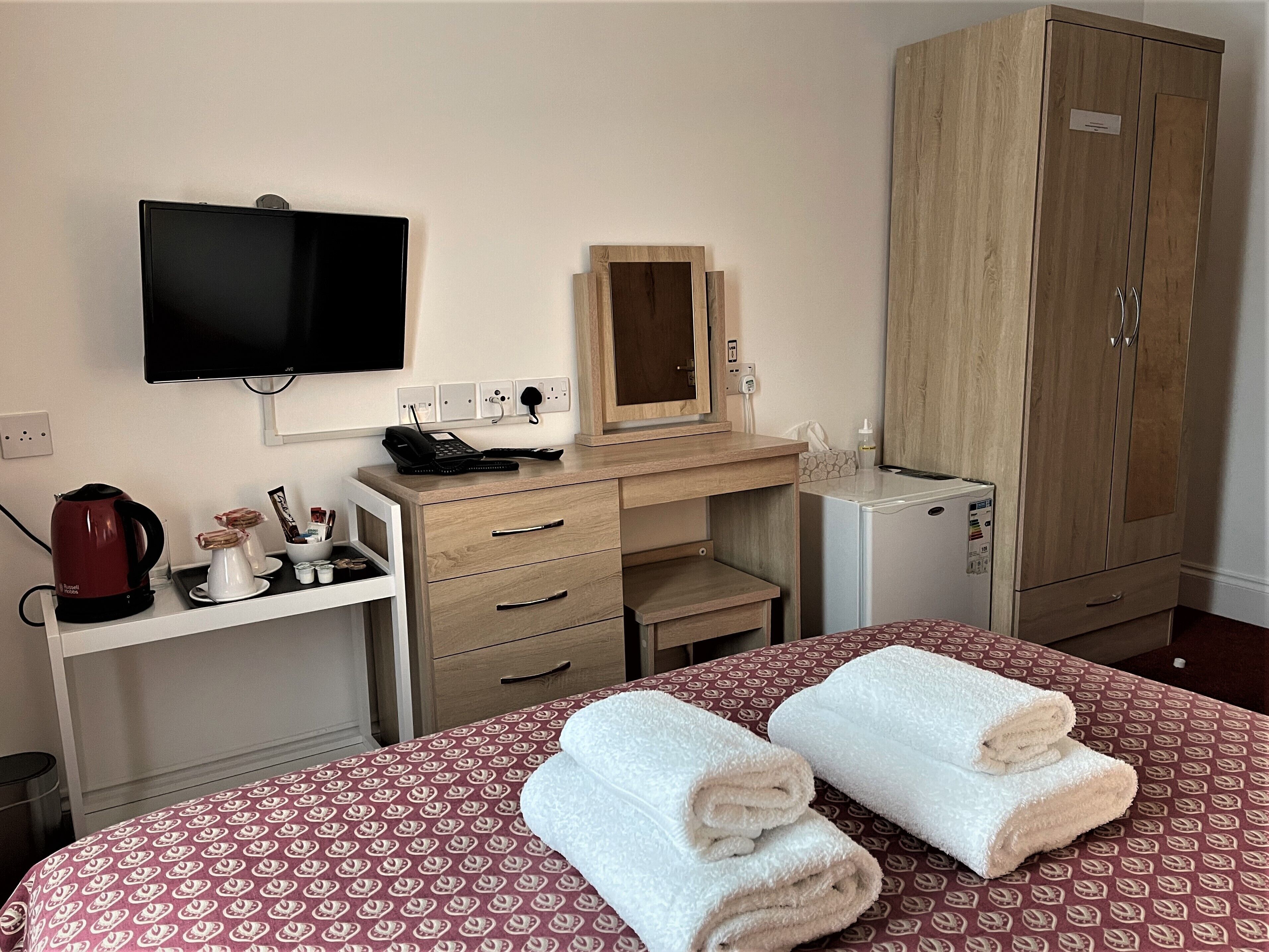 A hotel room with a bed, a desk with a telephone, a television, a wardrobe, and a small refrigerator.