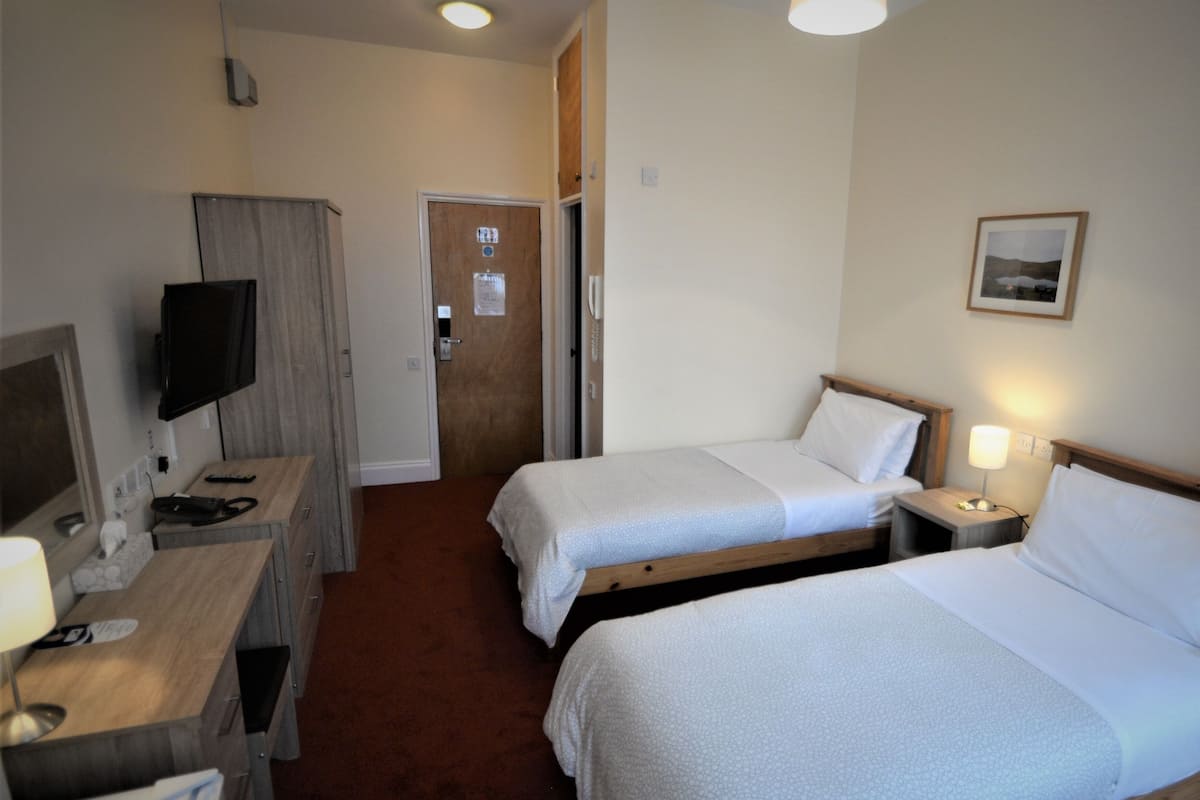 Quarto de hotel com duas camas de solteiro, uma escrivaninha com computador e televisão, uma porta e uma pequena mesa de cabeceira com abajur.