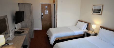 A hotel room with two single beds, a desk with a computer and a TV, a door, and a small bedside table with a lamp.