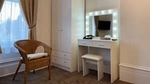 A room with a white vanity, a wicker chair, a white desk, and a large mirror with lights. - Gateway Hotel (London)