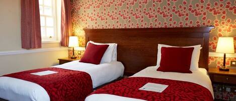 Habitación de hotel con dos camas individuales, cada una con una colcha roja y blanca y almohadas a juego. Hay dos mesitas de noche con lámparas, y el papel tapiz de la habitación tiene un estampado floral.