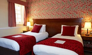 A hotel room with two single beds, each with a red and white bedspread and matching pillows. There are two bedside tables with lamps, and the room has floral wallpaper. - Saracens Head Towcester by Greene King Inns (Towcester)