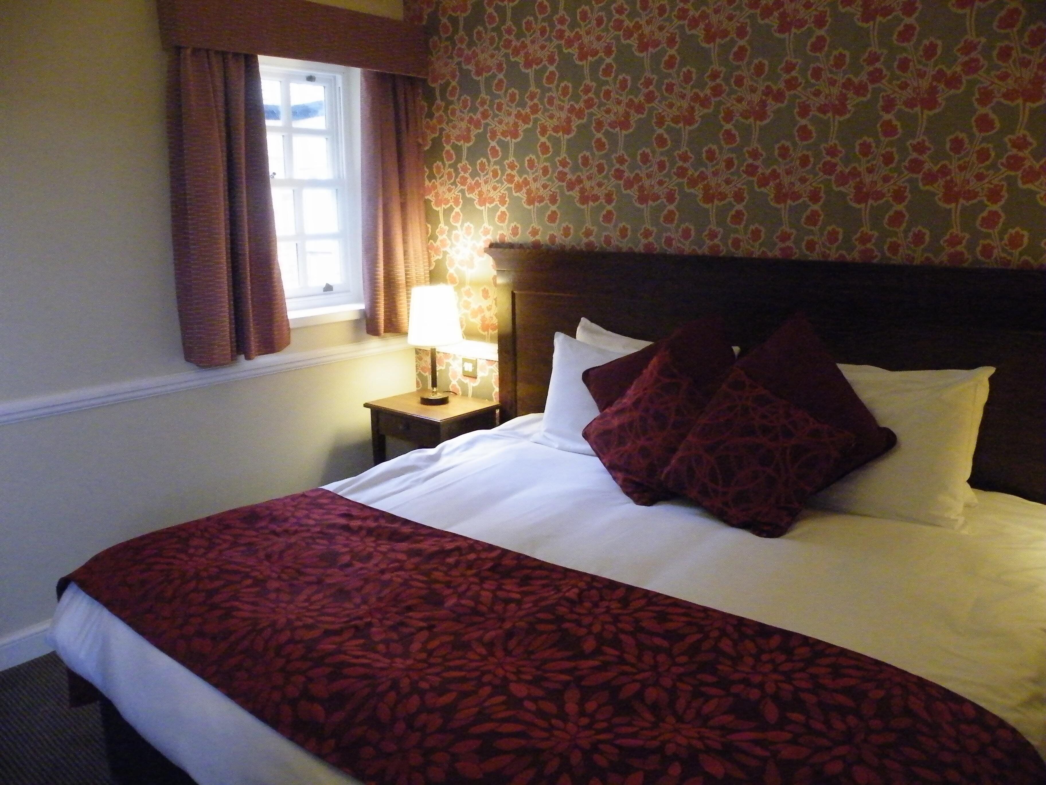A hotel room with a neatly made bed, a bedside table with a lamp, and a window with curtains.