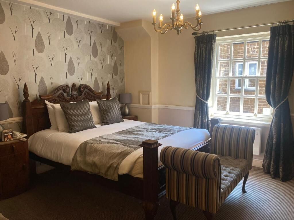 A hotel room with a large bed, a chair, a nightstand, a chandelier, and a window with curtains.