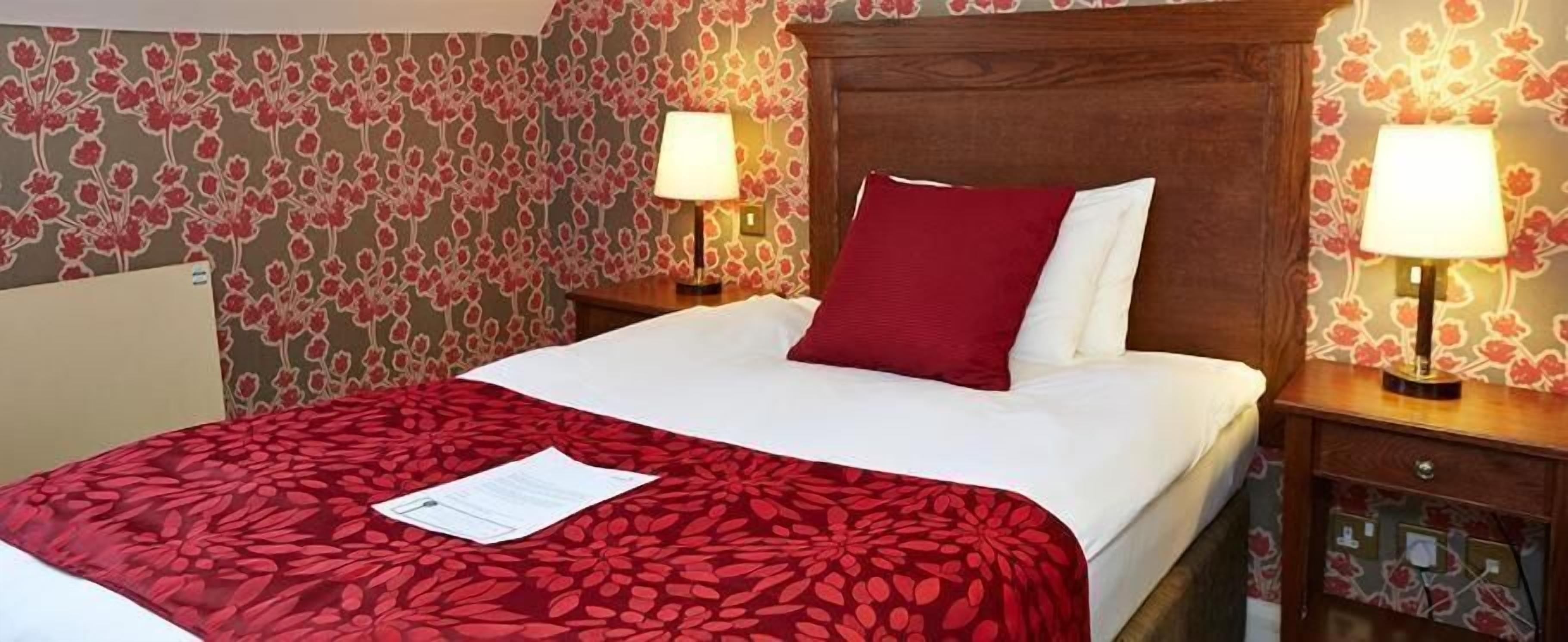 Habitación de hotel con cama, colcha roja estampada con flores, dos lámparas de noche y papel tapiz floral.