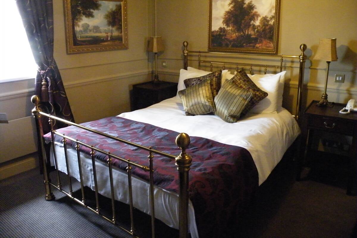 Una cama bien hecha con una colcha estampada, dos mesitas de noche con lámparas, y dos cuadros con paisajes en las paredes.