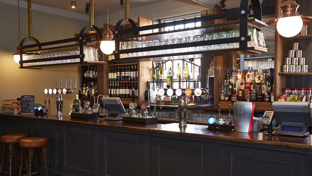 A bar area with multiple shelves of bottles, a cash register, and stools. - Saracens Head Towcester by Greene King Inns (Towcester)
