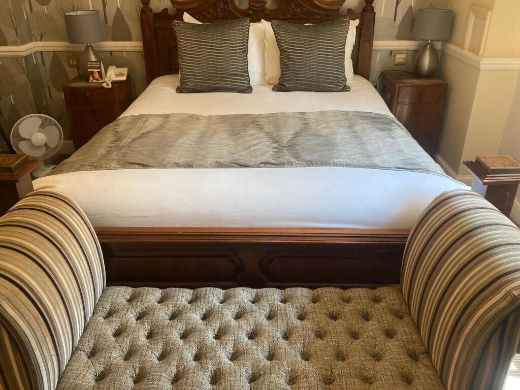 Una cama bien tendida con un cabecero capitonado, dos almohadas y una mesita de noche con una lámpara.