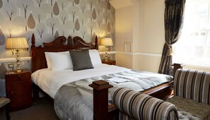 A hotel room with a large bed, two bedside lamps, a patterned wall, a window with curtains, and striped armchairs. - Saracens Head Towcester by Greene King Inns (Towcester)