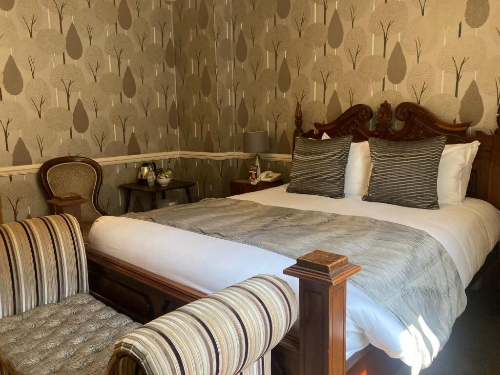 A hotel room with a large bed, two upholstered chairs, a nightstand with a lamp, and a patterned wallpaper.