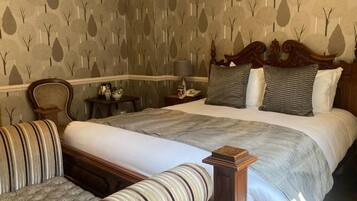 A hotel room with a large bed, two upholstered chairs, a nightstand with a lamp, and a patterned wallpaper.