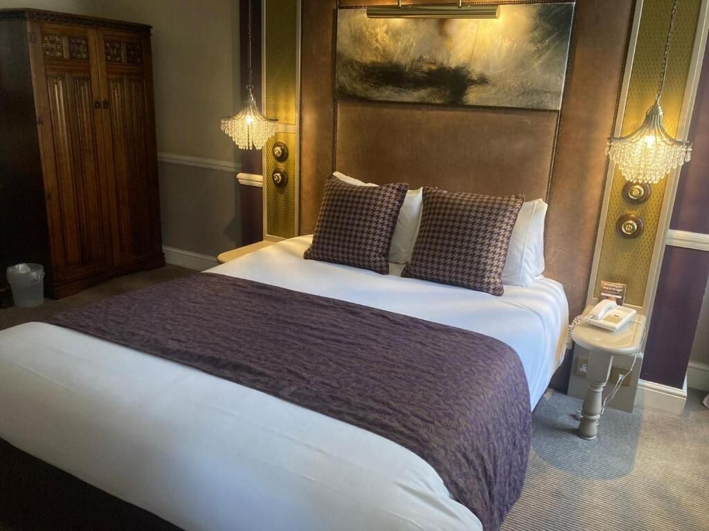 Habitación de hotel con una cama grande, cabecera con una pintura de paisaje, dos lámparas de noche y un armario de madera.