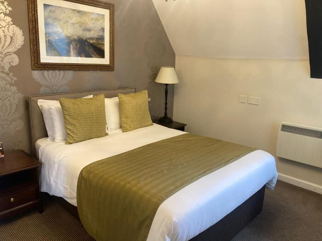 A hotel room with a bed, nightstand, lamp, and a framed picture on the wall.