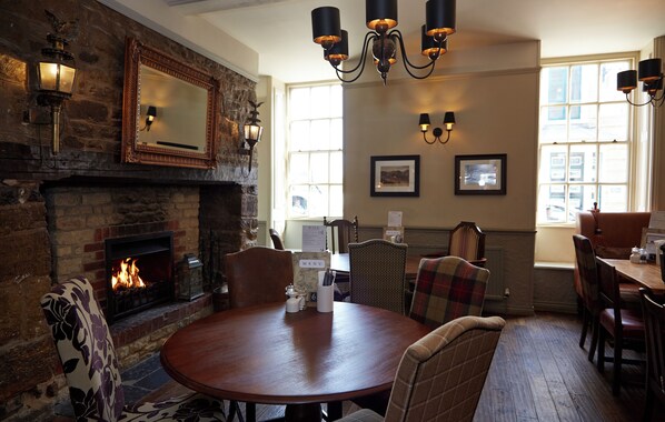 A cozy dining area with a fireplace, wooden table, and chairs. There are wall-mounted lights and a chandelier. - Saracens Head Towcester by Greene King Inns (Towcester)