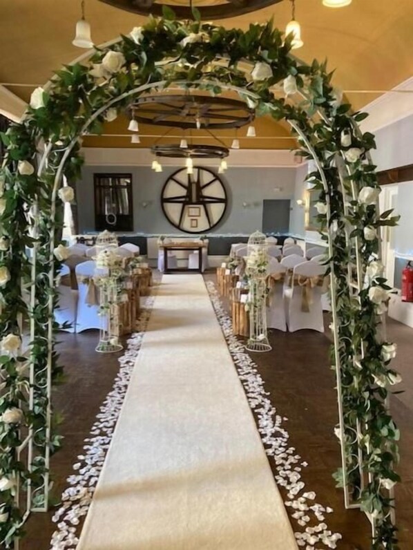 An indoor venue with a floral archway leading to a long table with chairs, surrounded by greenery and white flowers. - Saracens Head Towcester by Greene King Inns (Towcester)