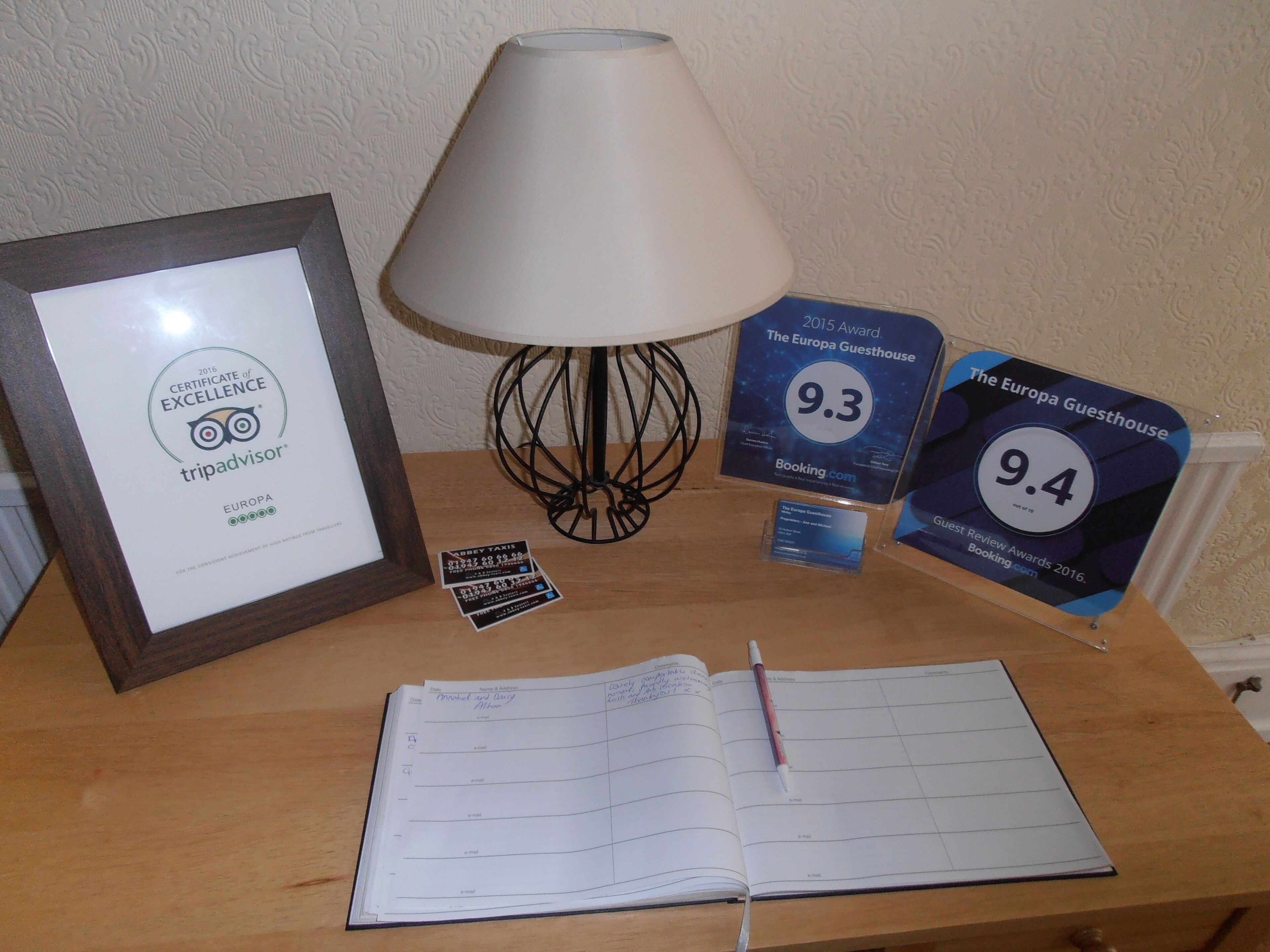 A wooden table with a framed certificate, a lamp, a notebook, and two guest review cards.