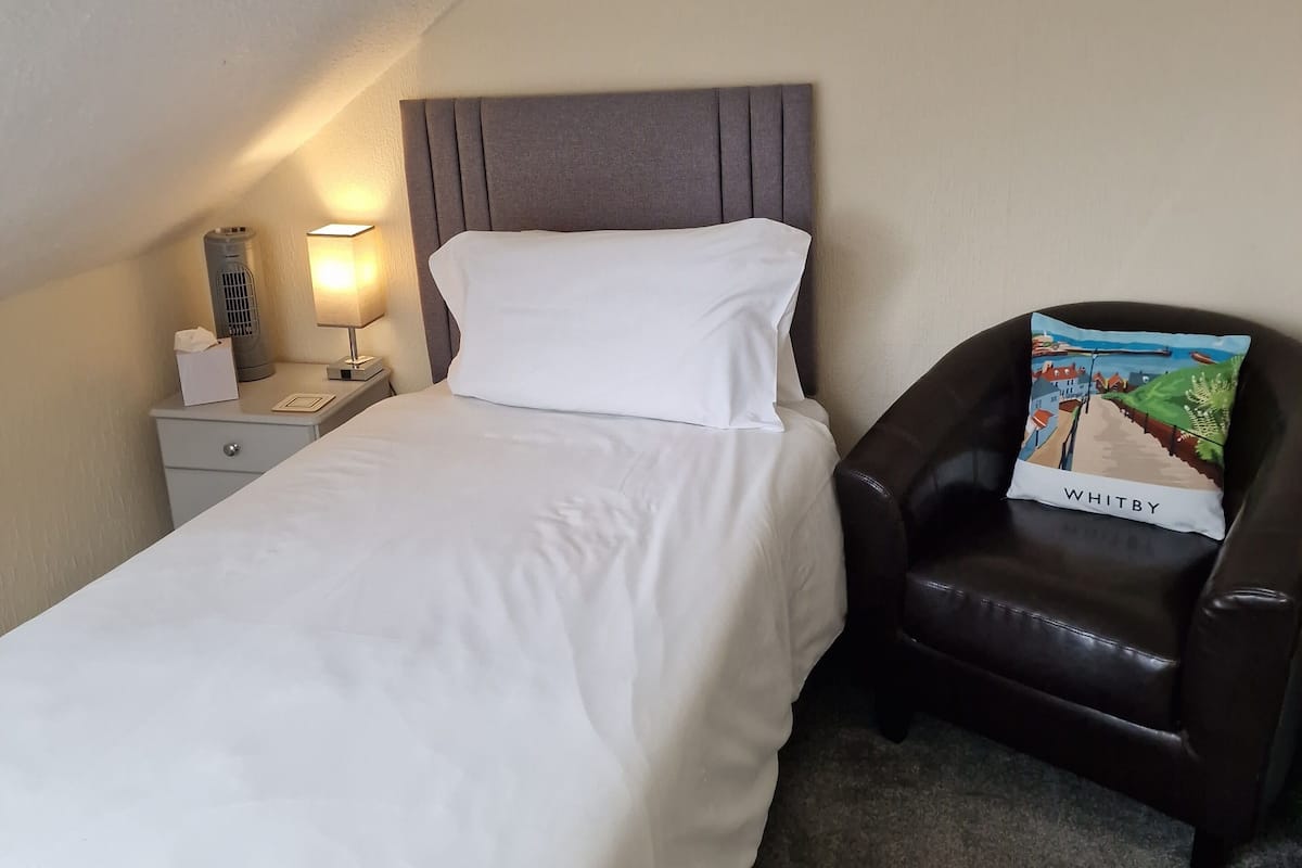 A hotel room with a bed, a nightstand with a lamp, a chair, and a pillow with the word "WHITBY".