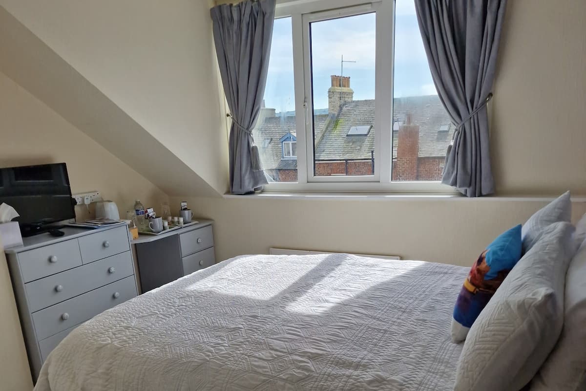A bedroom with a large bed, a desk with a computer, a window with curtains, and a view of buildings outside.