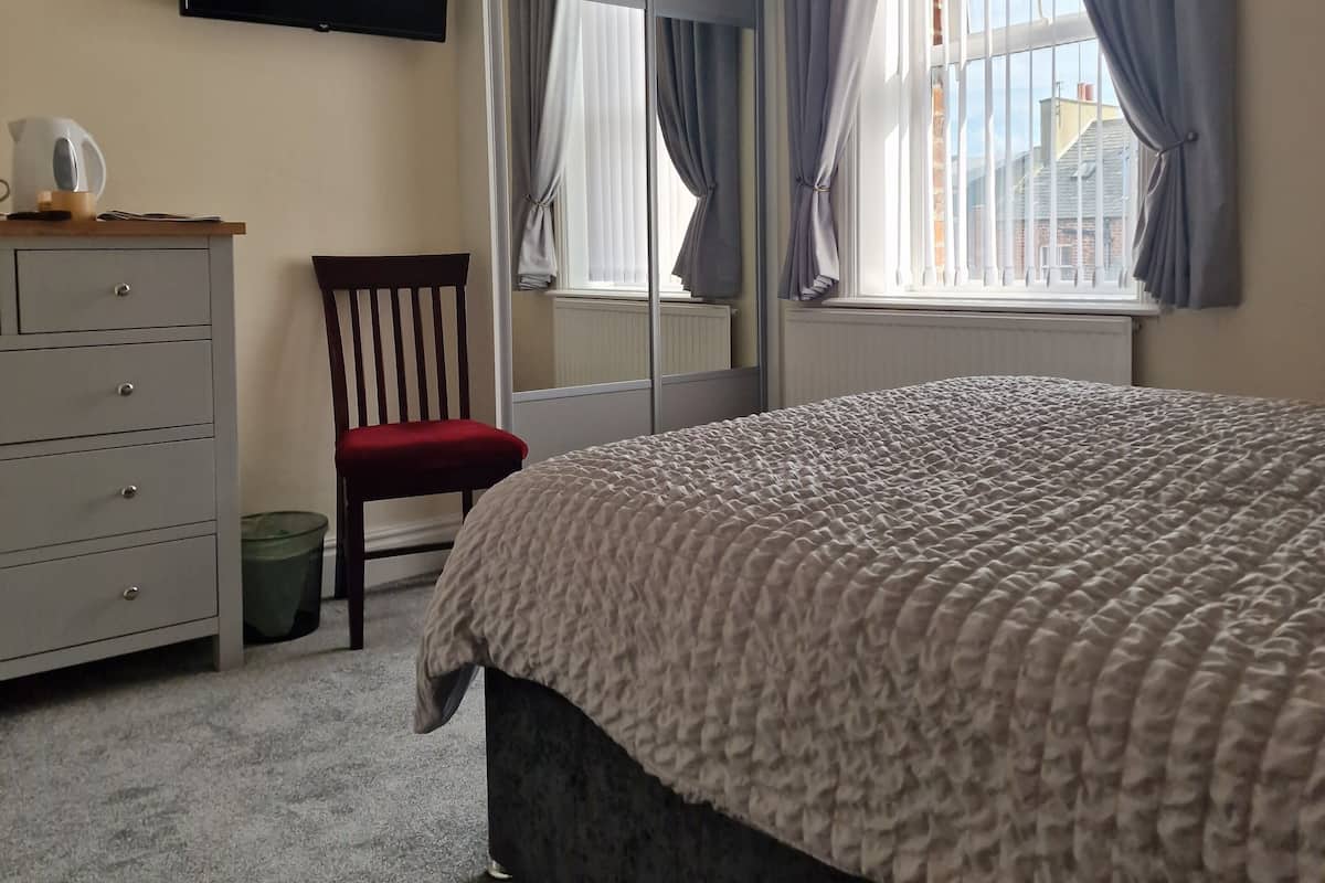 A bedroom with a bed, a TV mounted on the wall, a dresser, a chair, and a window with curtains.