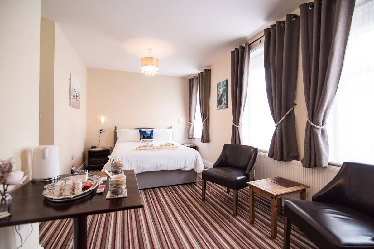 A hotel room with a bed, a desk with a tray of breakfast items, a chair, a small table, and a window with curtains.