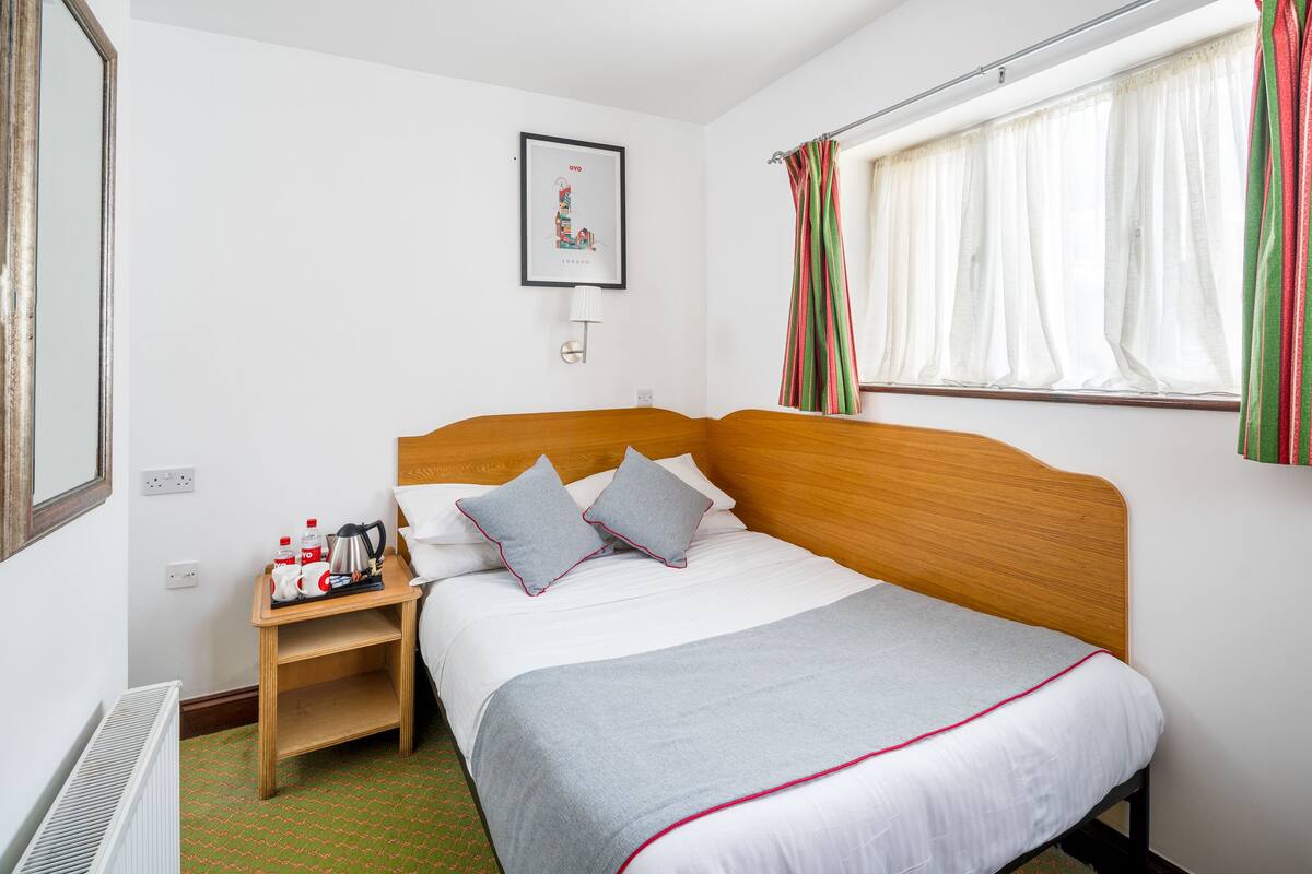 A neatly made bed with a grey blanket and pillows, a bedside table with a kettle, and a framed picture on the wall.