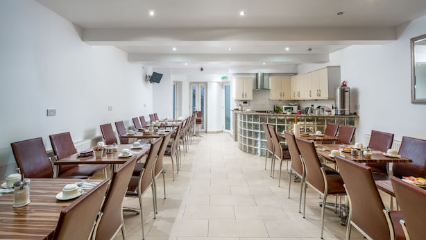 A dining area with multiple tables and chairs, a kitchen in the background, and a tiled floor. - Abbey Hotel Shepherd’s Bush (London)