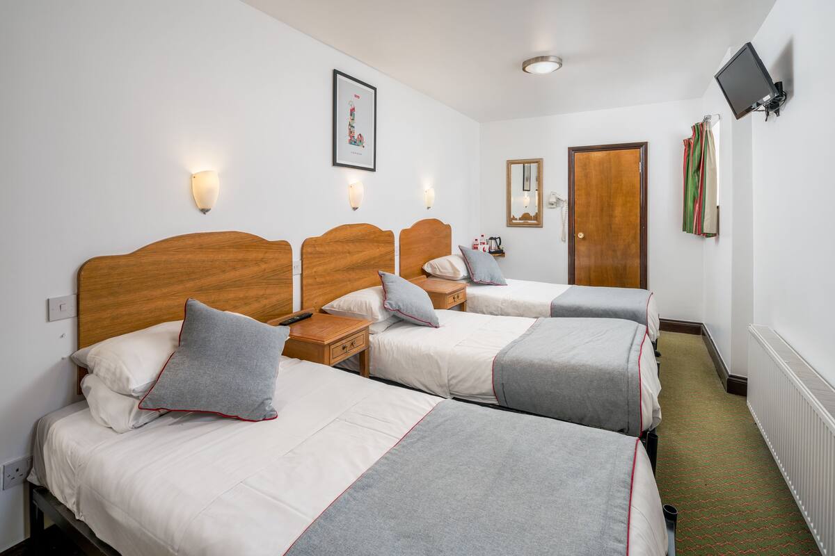 A hotel room with three single beds, wooden headboards, bedside tables, a flat-screen TV mounted on the wall, and a framed picture on the wall.