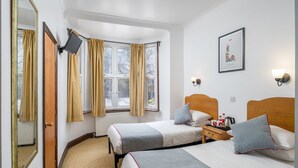 A hotel room with two beds, a wooden headboard, a nightstand with a lamp, a framed picture on the wall, and a window with curtains. - Abbey Hotel Shepherd’s Bush (London)
