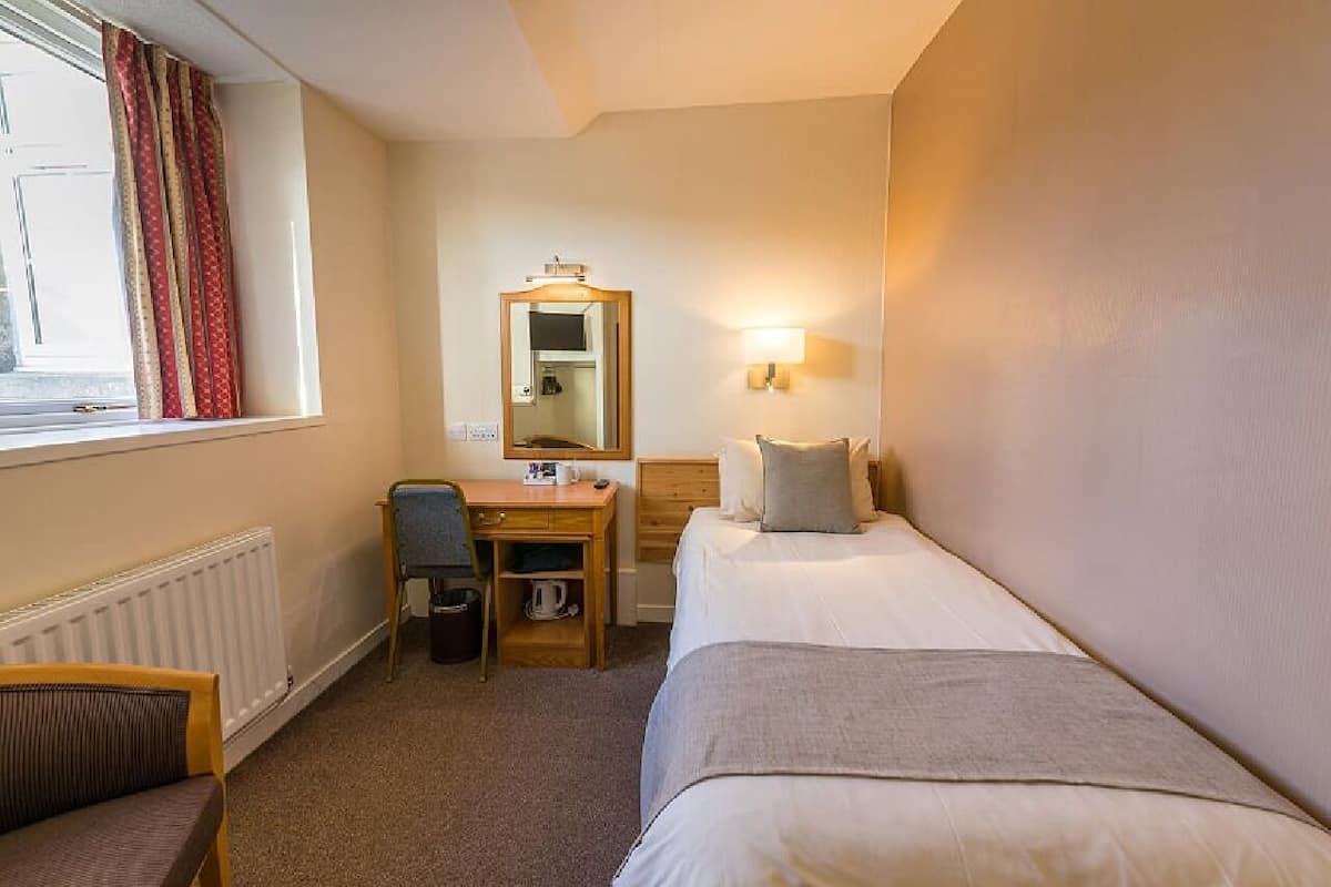 A hotel room with a single bed, a desk with a chair, a mirror, and a window with curtains.
