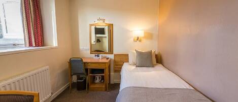 Quarto de hotel com uma cama de solteiro, uma escrivaninha com cadeira, um espelho e uma janela com cortinas.