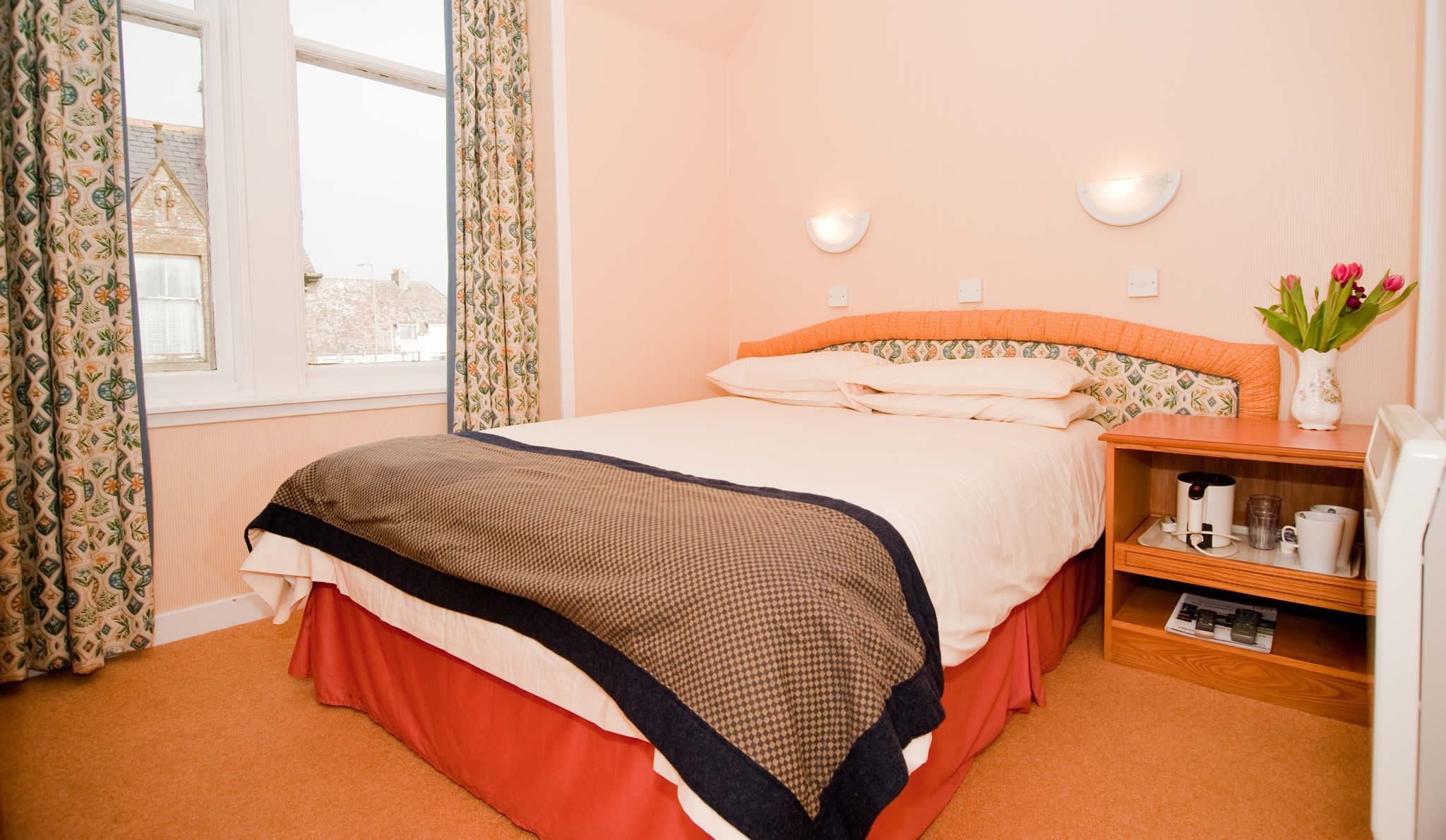 A neatly made bed with a patterned headboard, a bedside table with a vase of flowers, and a window with patterned curtains.