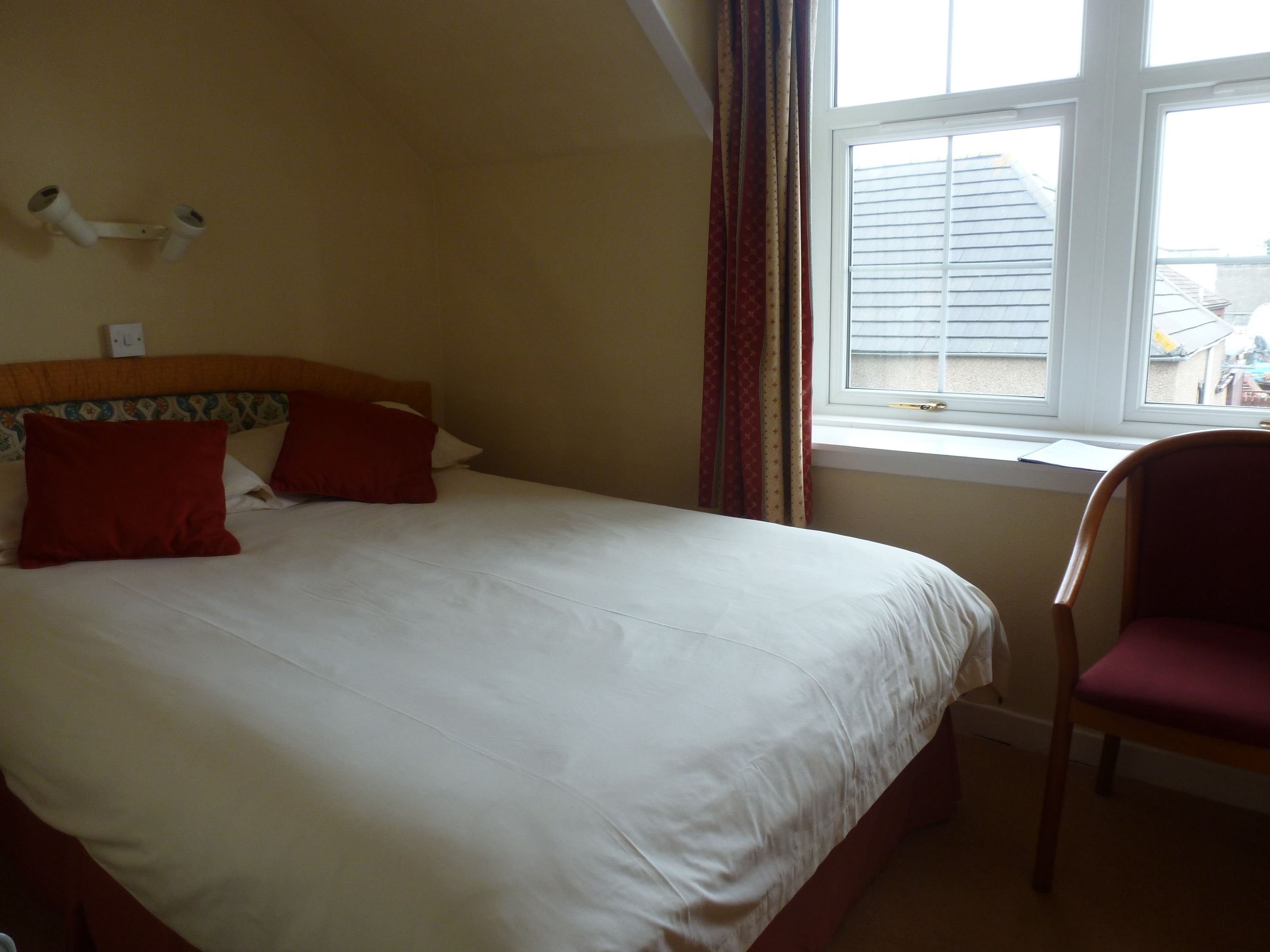 A bedroom with a bed, a chair, a window with blinds, and a wall-mounted light switch.