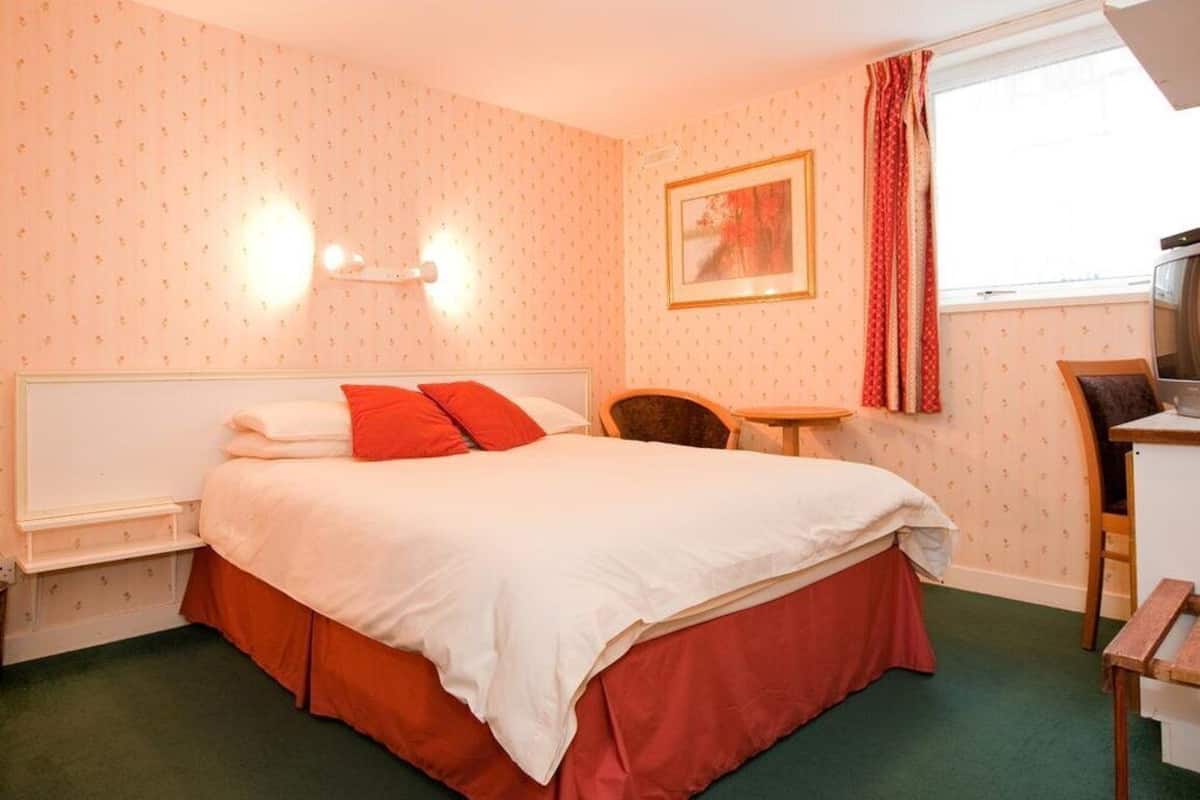 A bedroom with a bed, a desk, a chair, a window with curtains, and a wall-mounted picture.