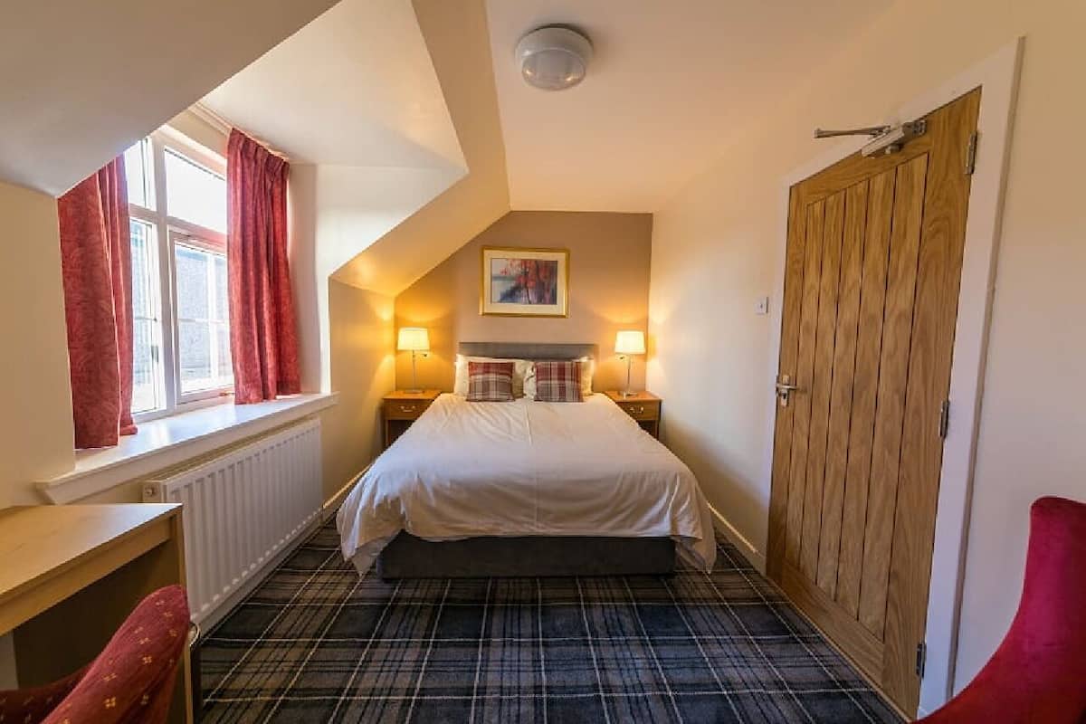 A bedroom with a bed, two bedside lamps, a window with curtains, a radiator, a wooden door, and a red chair.