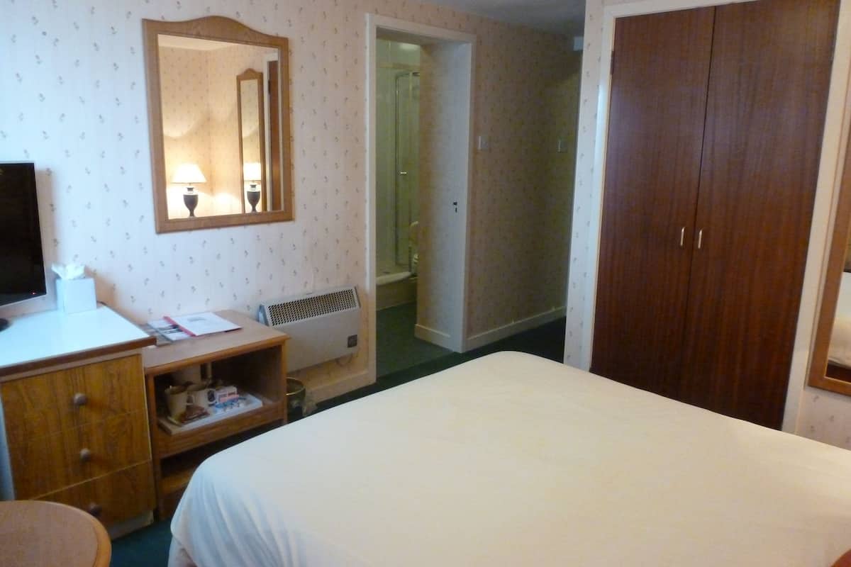 A hotel room with a bed, a desk with a lamp, a mirror, a wardrobe, and a bathroom visible through an open door.
