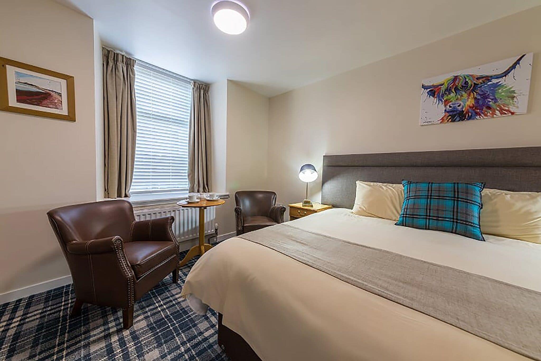 A hotel room with a large bed, a headboard, a desk with a chair, a window with curtains, and a painting on the wall.