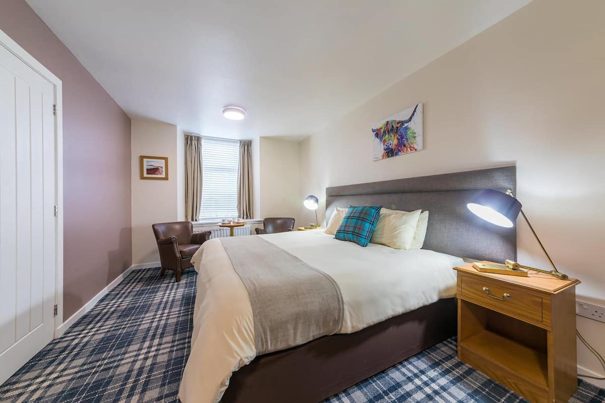 A hotel room with a bed, bedside table, lamp, chair, and a window with curtains.