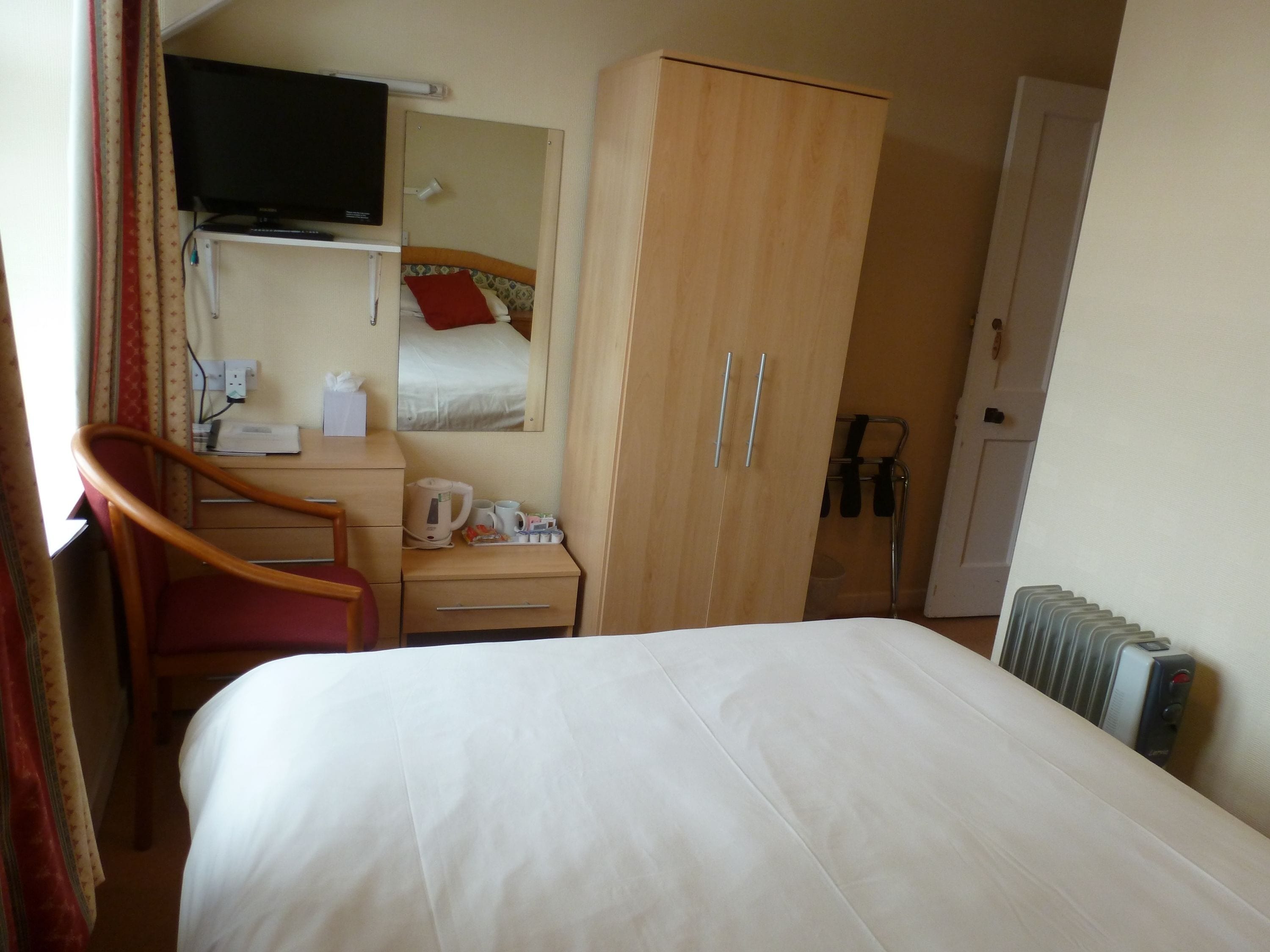 Una habitación de hotel con cama, escritorio y silla, armario, televisor montado en la pared y radiador.