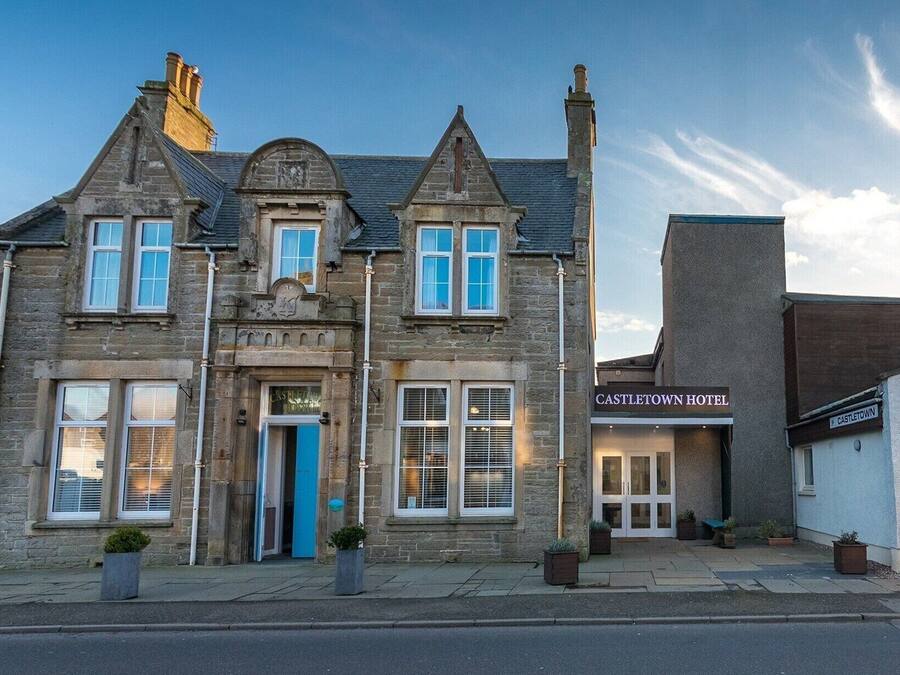 Un bâtiment historique en pierre, de style architectural traditionnel, qui se distingue par son entrée imposante avec une porte bleue, de nombreuses fenêtres et une enseigne indiquant l’hôtel Castleletown.