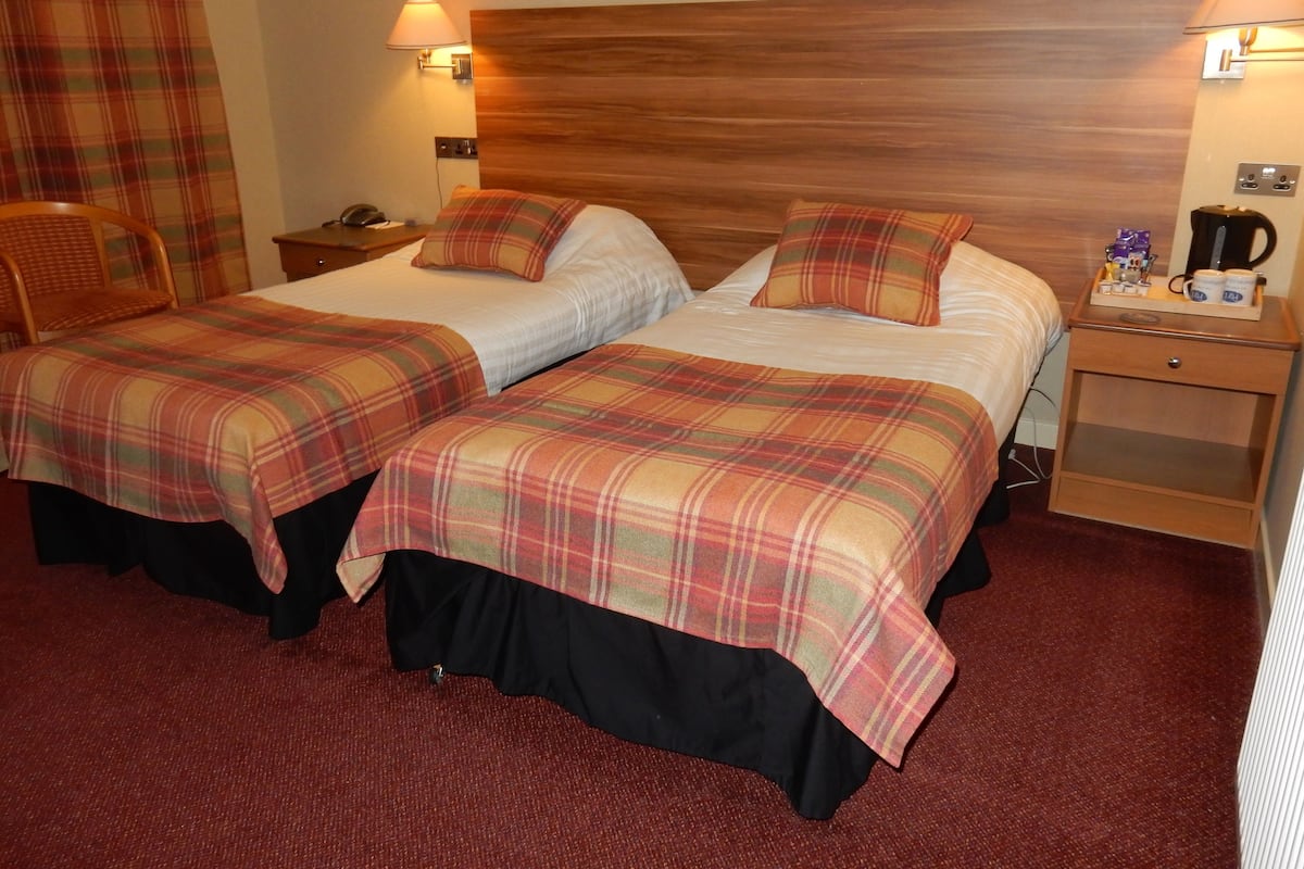 A hotel room with two beds, each with a plaid bedspread, a wooden headboard, bedside lamps, and a nightstand with a coffee maker.