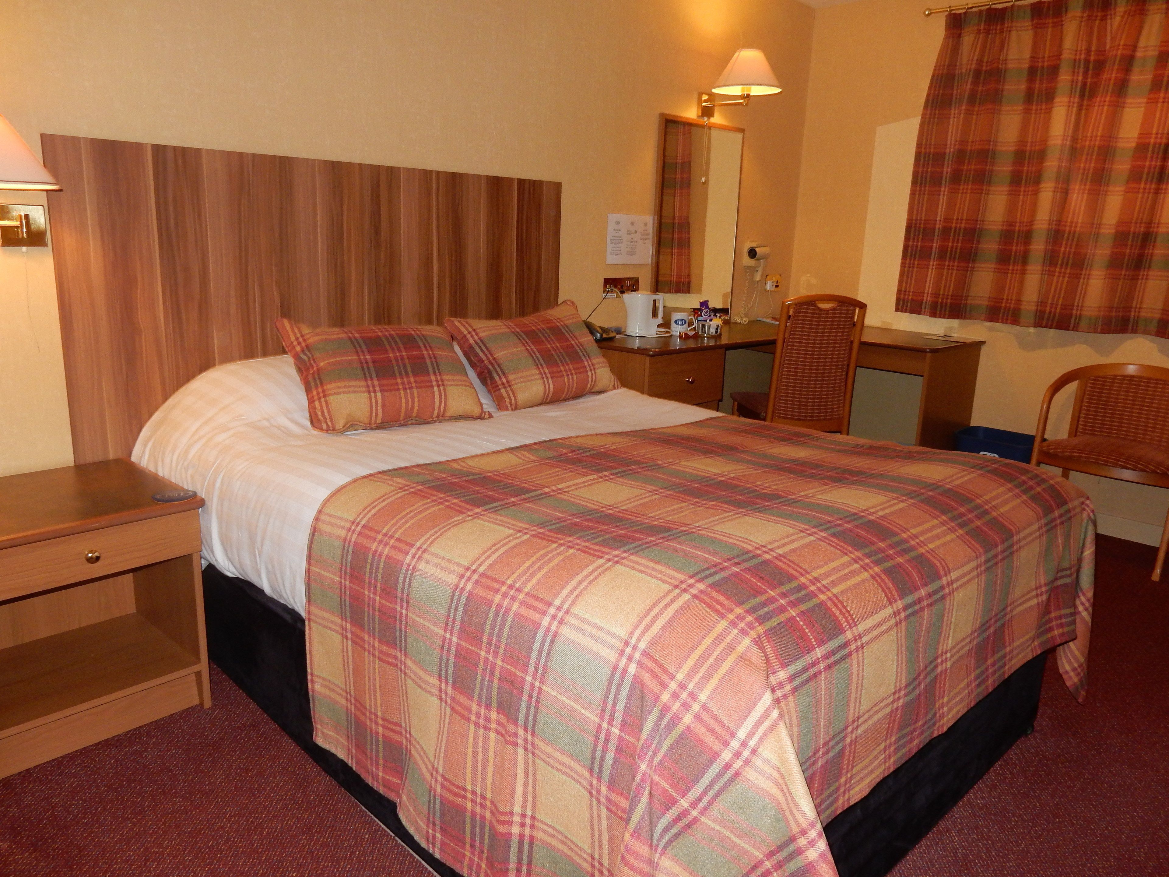 A hotel room with a large bed, a wooden headboard, a desk with a chair, a mirror, and a window with curtains.