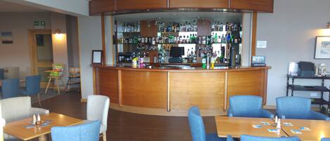 A bar area with a wooden counter and shelves of various liquor bottles. There are blue upholstered chairs and wooden tables with menus.