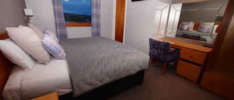 A bedroom with a bed, a desk, a chair, a mirror, a TV, and a window with curtains.